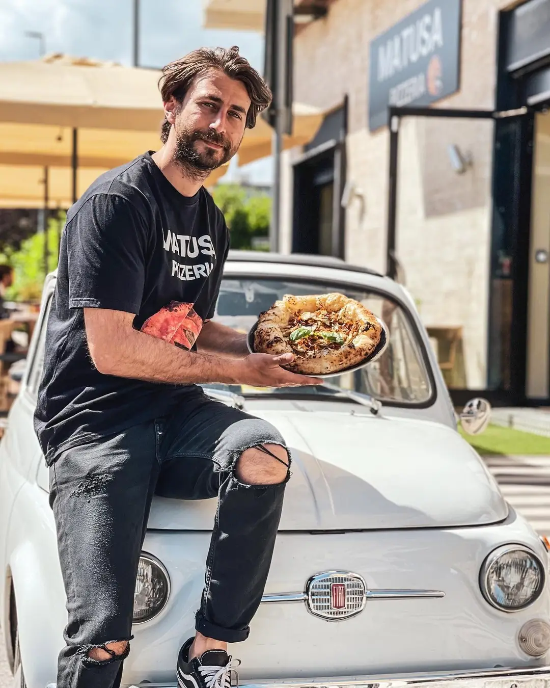 Adriano Russo con una pizza in mano, seduto su una Fiat 500 davanti all'insegna di Matusa